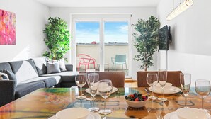 Ein Essbereich mit einem Holztisch, gedeckt für vier Personen, umgeben von einer Couch und Sesseln, mit Balkonblick im Hintergrund.