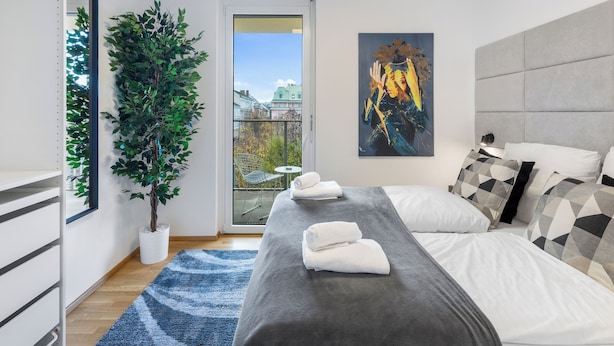 Ein modernes Schlafzimmer mit einem Bett, einer großen Topfpflanze, einem Gemälde und Blick auf die Stadt durch eine Glastür.