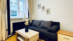 Una habitación con un sofá, una pequeña mesa con un jarrón de flores, una ventana con cortinas y tres cuadros enmarcados en la pared.