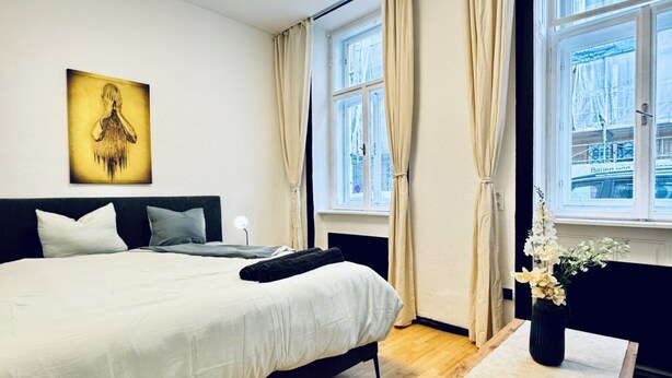 Un dormitorio con una cama grande, una mesita de noche con un jarrón de flores, un cuadro en la pared y una ventana con cortinas.