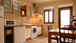 Une cuisine compacte équipée d’un lave-vaisselle, d’un lave-linge et d’un four à micro-ondes. On y trouve une table à manger avec des chaises et une fenêtre avec des stores.