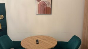 Une pièce comprenant une table ronde en bois, deux chaises de couleur turquoise, une œuvre d’art abstraite encadrée accrochée au mur et un petit objet noir posé sur la table.