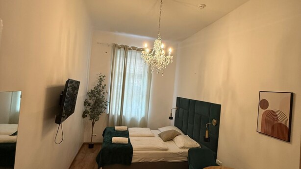 Une chambre d’hôtel avec un lit, un bureau, une chaise, une table ronde, un lustre, une plante et un tableau au mur.