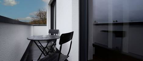 Ein Balkon im Freien mit Tisch und Stühlen, einer weißen Wand und Blick auf Himmel und Bäume.