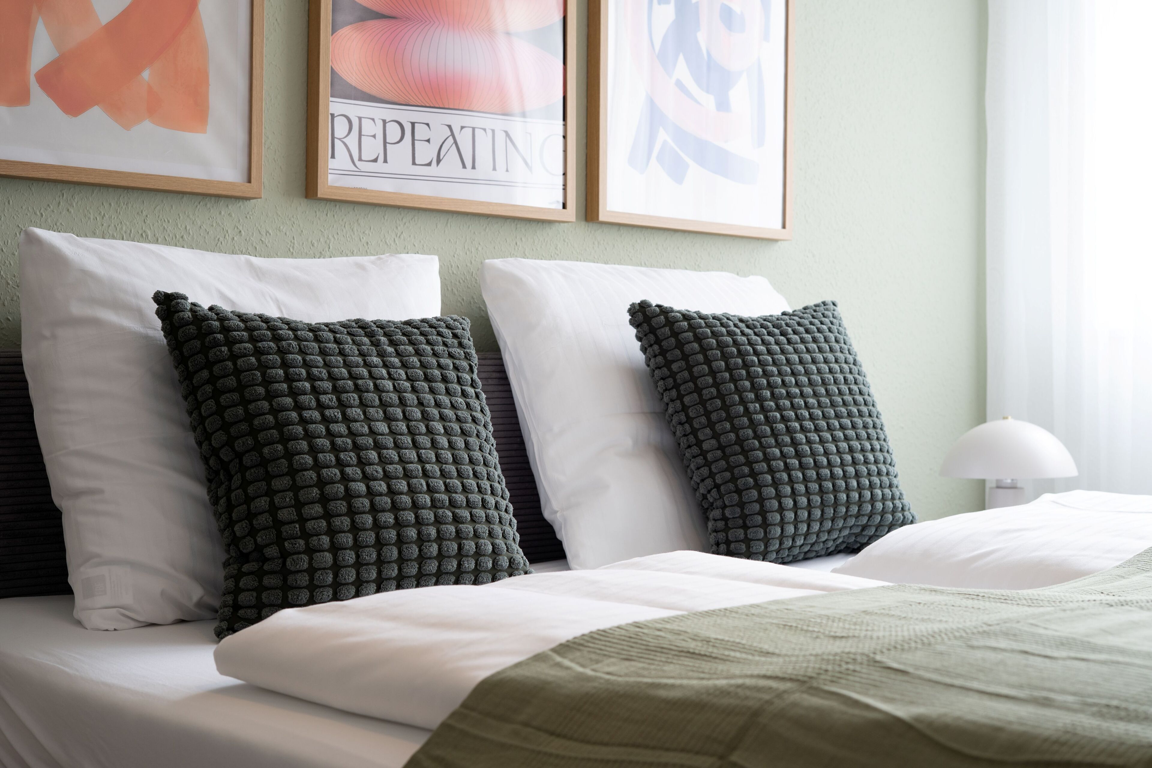A neatly made bed with two dark green textured pillows and a white pillow, set against a light green wall with framed artwork above.