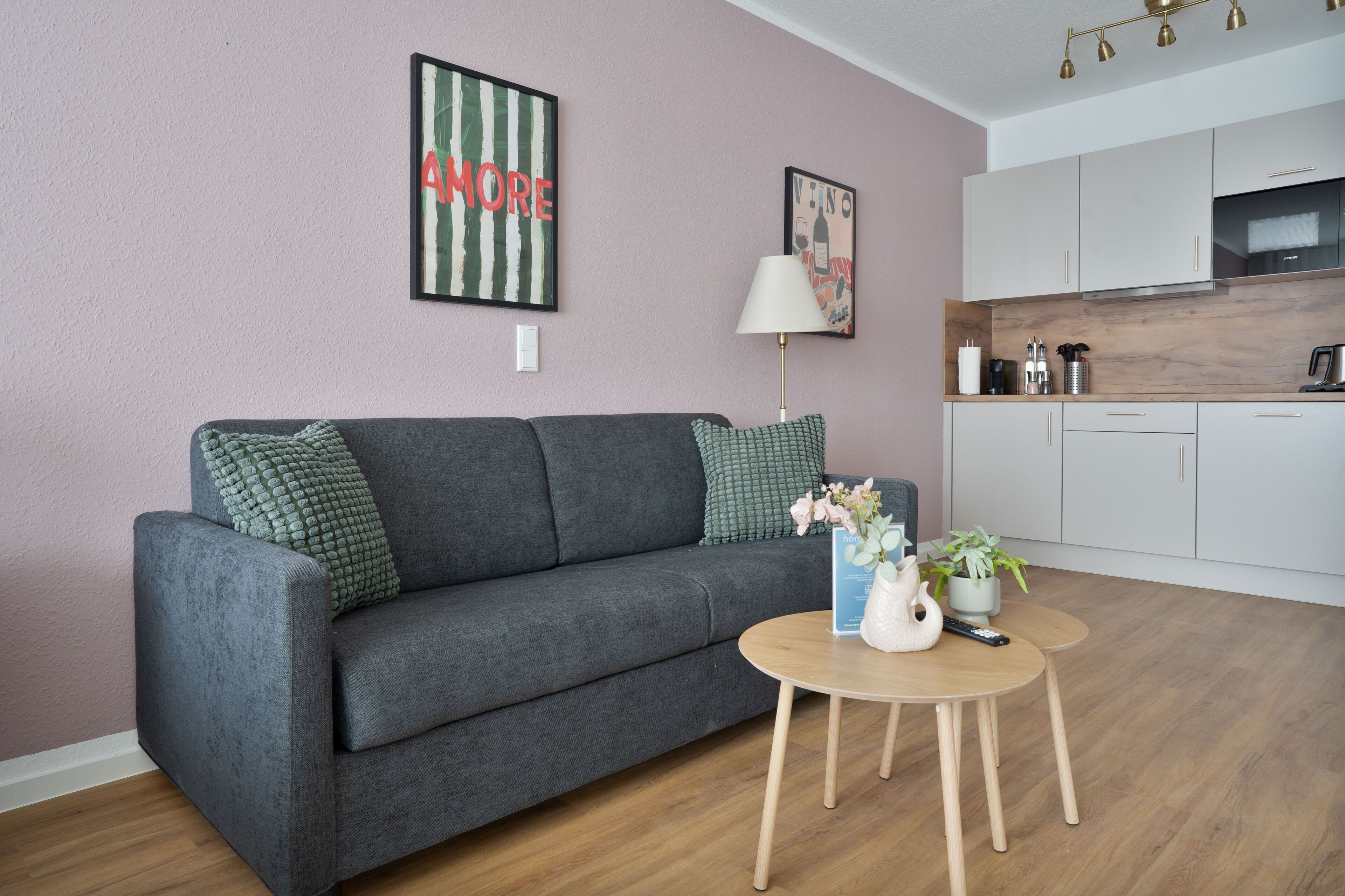 A modern living room with a grey sofa, a small round wooden coffee table, and a kitchen in the background.