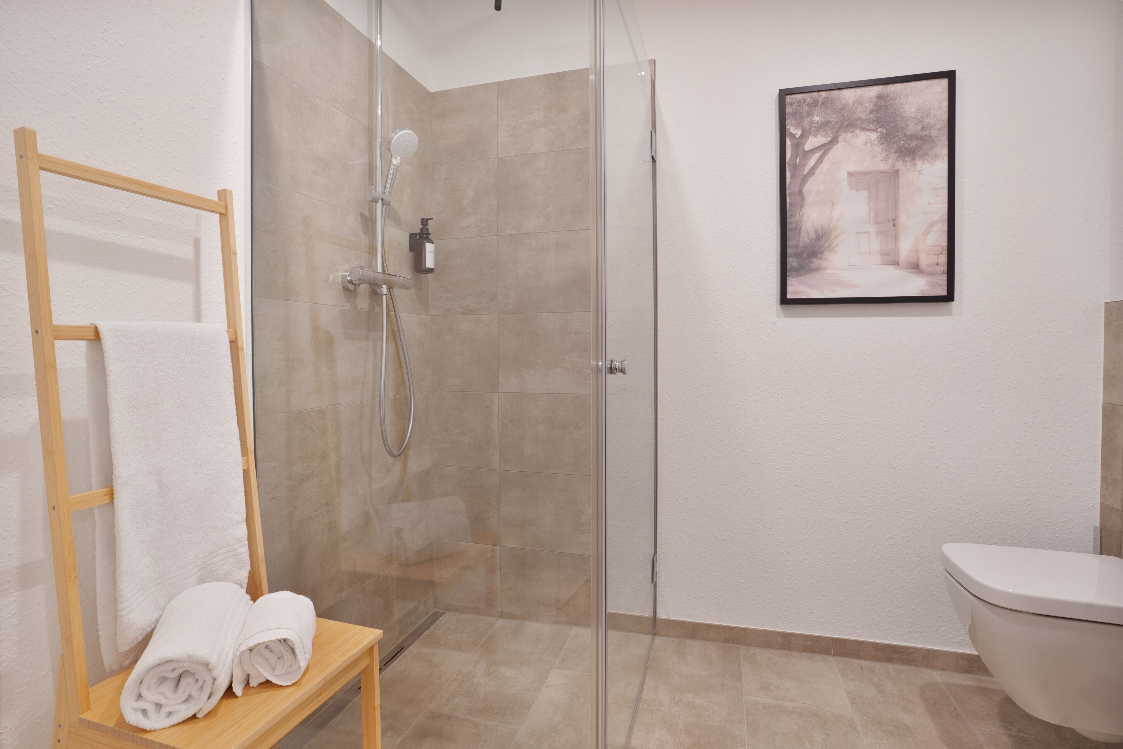 A modern bathroom with a glass shower enclosure, a wooden towel rack, and a framed picture on the wall.