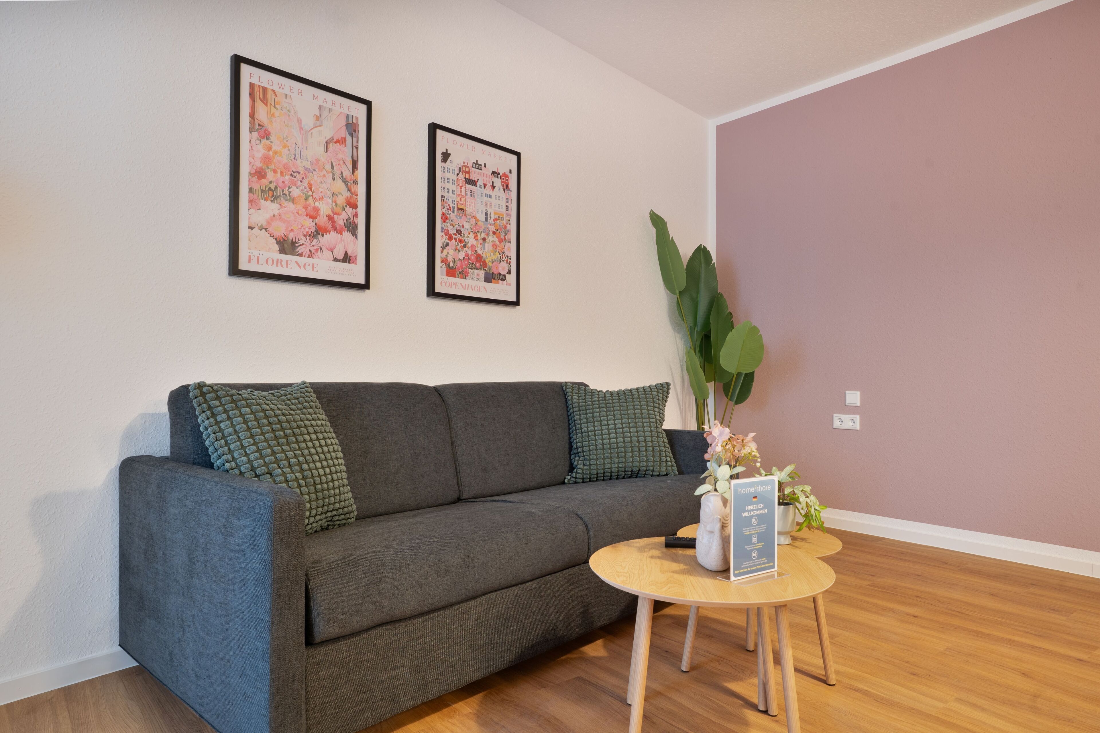 Una sala moderna con un sofá gris, dos carteles enmarcados con motivos florales, una planta verde y una pequeña mesa de madera con un letrero.