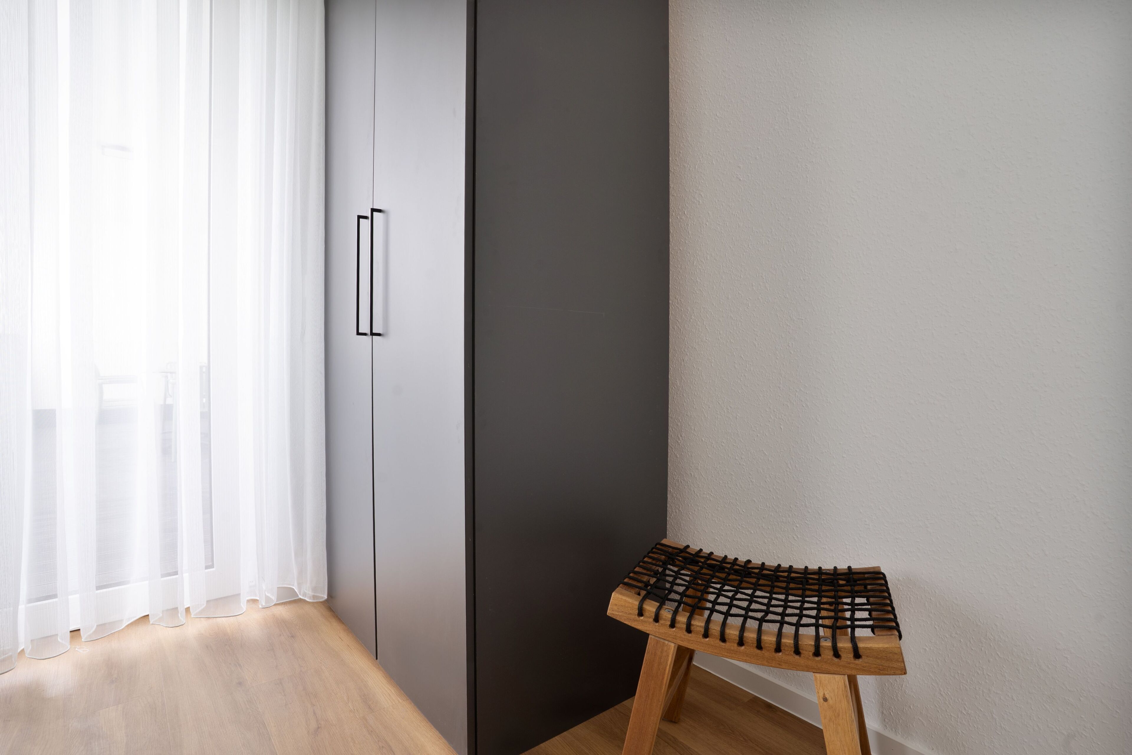 Ein Eckbereich eines Raumes mit Holzboden, einem schwarzen Kleiderschrank, einer weißen Wand und einem kleinen Holzschemel.