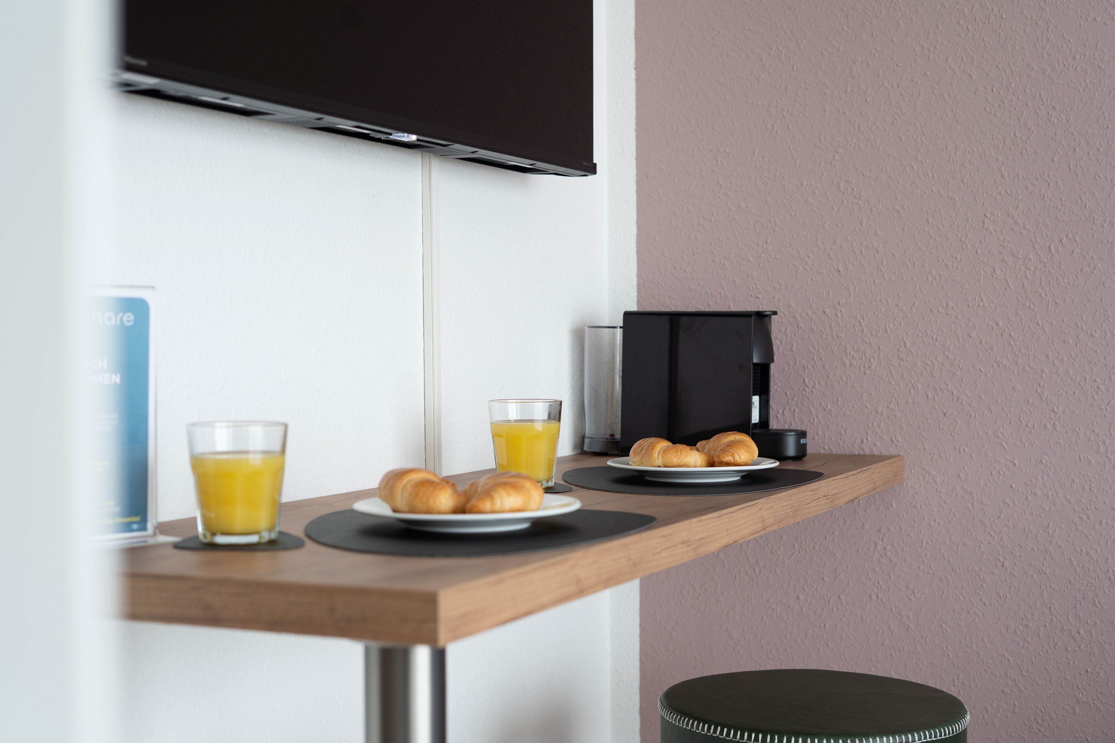 A breakfast setup with croissants, orange juice, and a coffee machine on a wooden table in a room with a TV mounted on the wall.