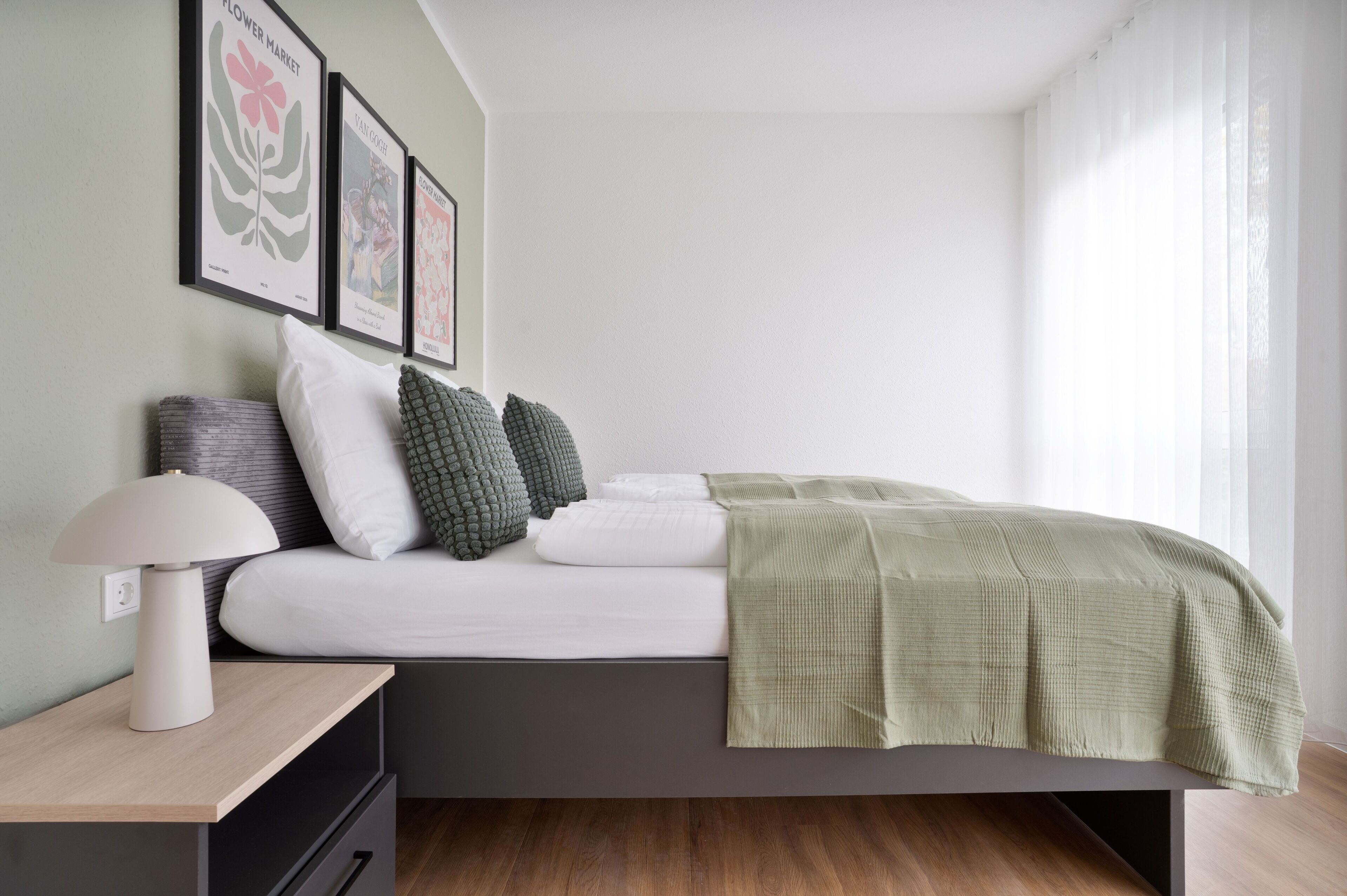 Ein Schlafzimmer mit einem Bett, Kissen, einem Nachttisch mit Lampe und gerahmten Bildern an der Wand.