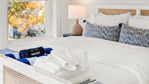 Ein ordentlich bezogenes Bett mit weißen Leinentüchern, ein blaues Handtuch mit der Aufschrift "MAKEUP" und ein weißes Handtuch. Neben dem Bett steht ein Nachttisch mit einer Lampe und einem Korbwäscheschrank.