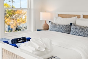 A neatly made bed with white linens, a blue towel labeled "MAKEUP," and a white towel. There is a wooden nightstand with a lamp and a wicker basket beside the bed.
