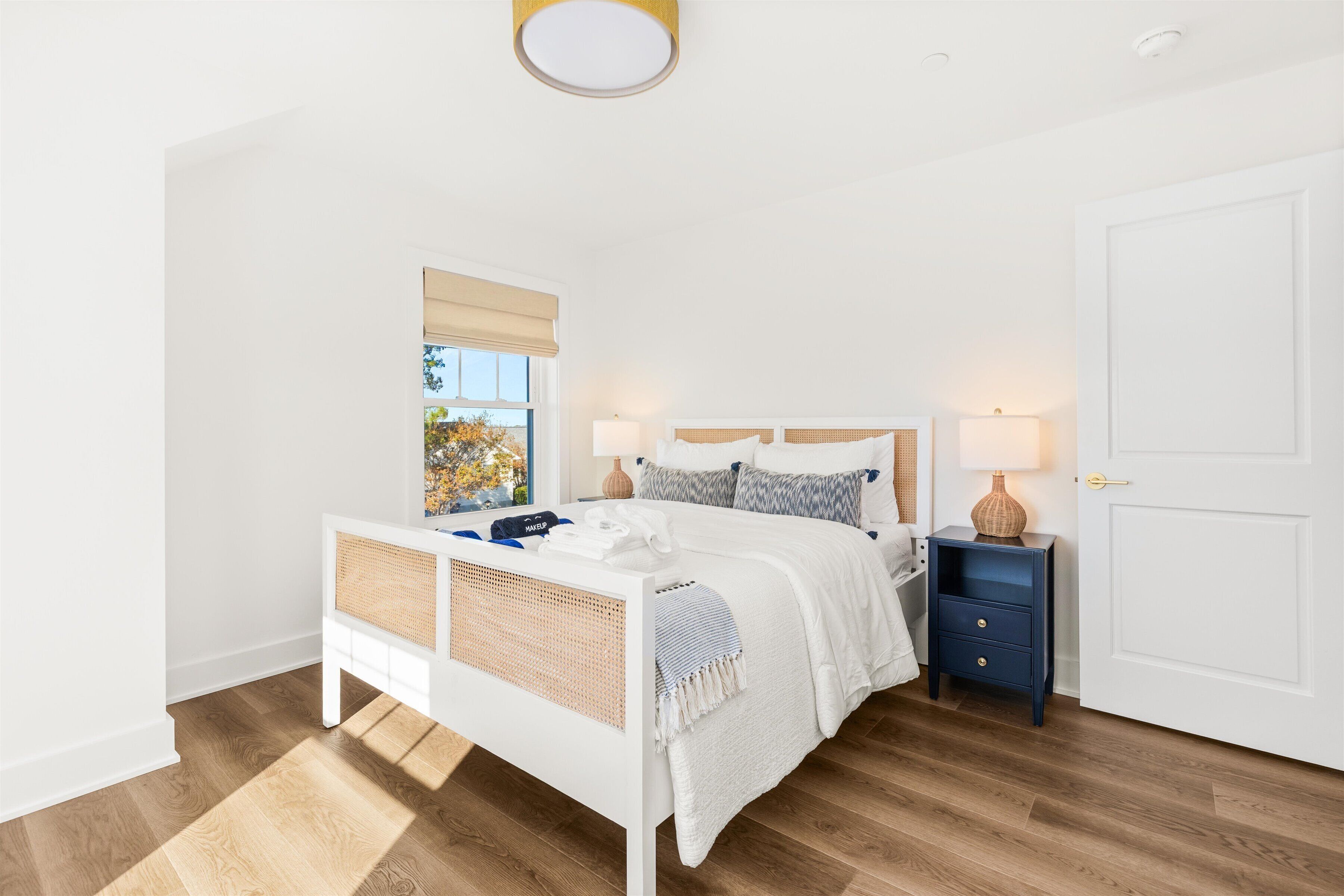 Een slaapkamer met een wit bed, een nachtkastje, een lamp en een raam met uitzicht op bomen.
