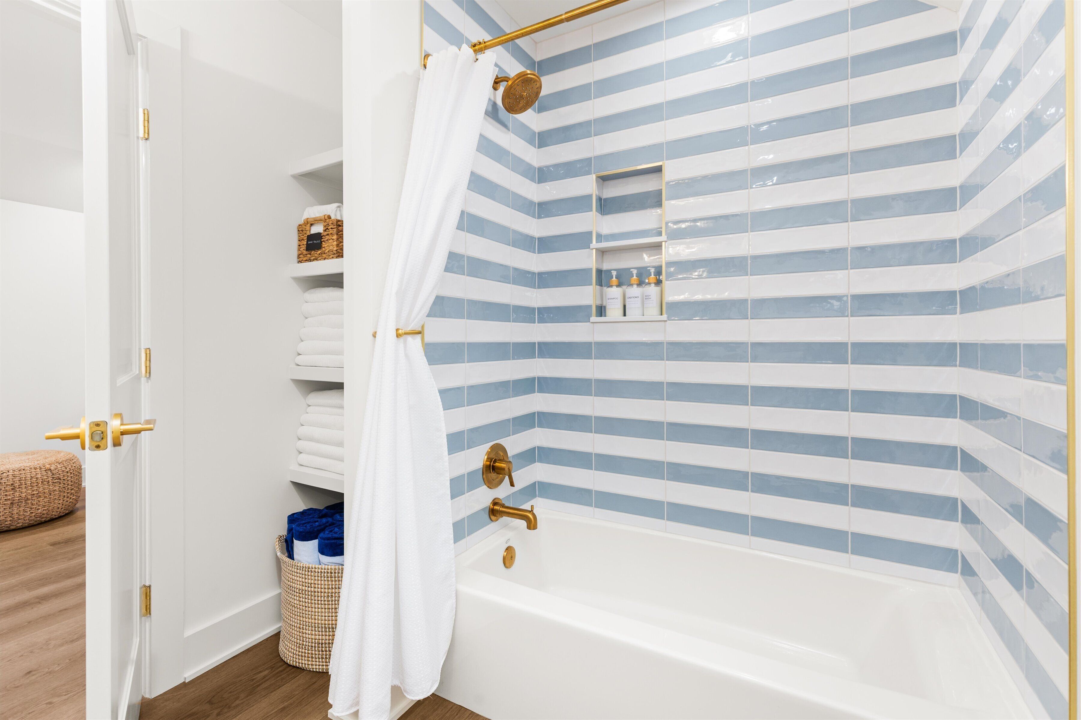 A bathroom with a white bathtub, blue and white striped tiles, a gold showerhead, and a towel rack with folded towels.