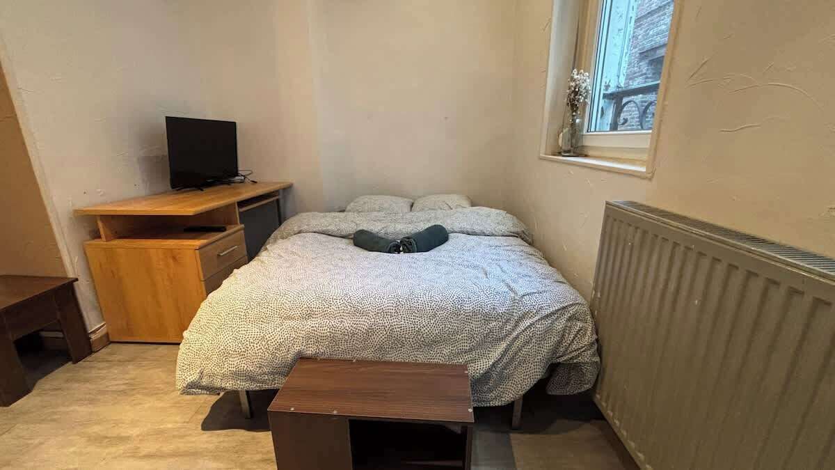 A small, sparsely furnished room with a bed, a wooden nightstand, a television, and a window with a view of buildings.