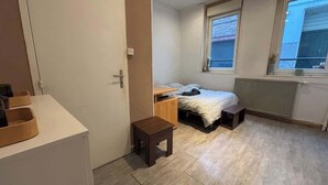 A compact bedroom with a single bed, a small wooden nightstand, a bench, and a radiator. - Petit rez-de-chaussée du théâtre (Arras)