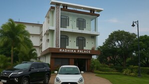 Un edificio de varios pisos llamado Radhika Palace con un diseño arquitectónico moderno, destacándose una entrada imponente y un jardín bien cuidado.
