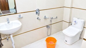 A bathroom with a white sink, a toilet, and a shower. There is a bucket on the floor. - Minx Hotels Radhika Palace (Ayodhya)