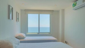A bedroom with two beds, a large window with a view of the sea, and an air conditioning unit on the wall.