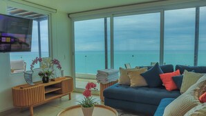 A living room with a large window overlooking the sea, a sofa with cushions, a wooden TV stand, and a small table with a plant.