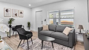 Ein modernes Wohnzimmer mit einem grauen Sofa, einem schwarzen Sessel und einem kleinen runden Tisch. An der Wand befinden sich gerahmte Kunstwerke, eine Tür und ein Fenster mit Jalousien.