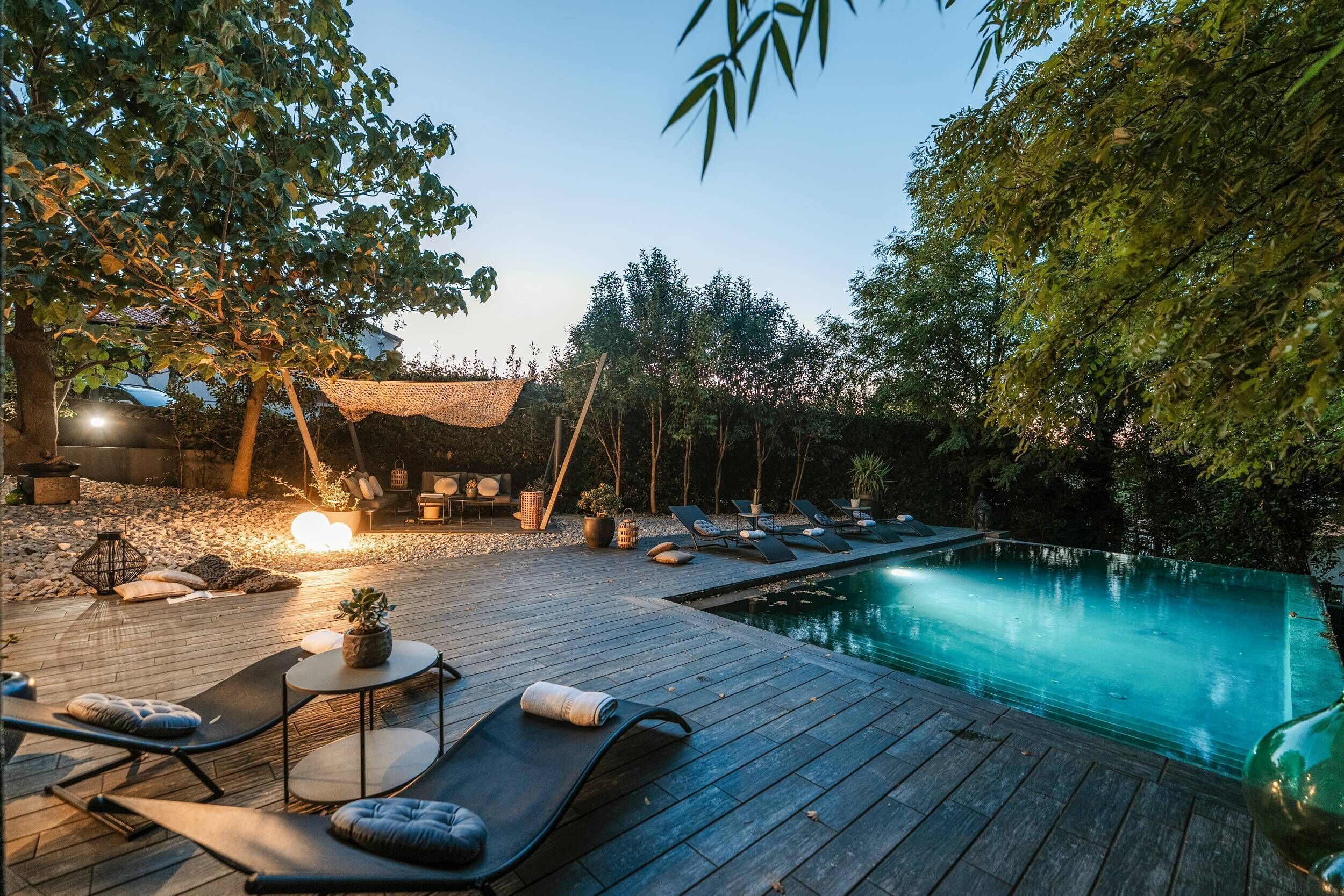 Una terraza de madera con sillones de descanso y una mesita, una piscina y árboles circundantes.