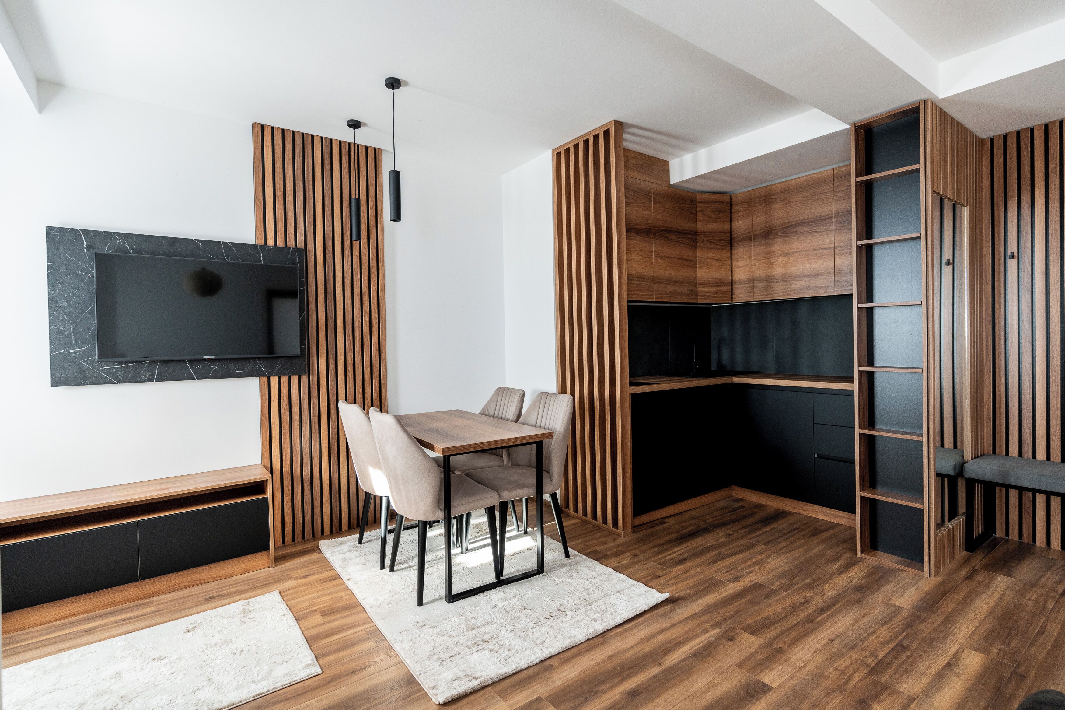 A modern living room with a wooden accent wall, a flat-screen TV mounted on the wall, a dining table with chairs, and a built-in shelving unit.