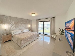 A modern bedroom with a large bed, a nightstand, a wall-mounted lamp, a sliding glass door to a balcony, and a painting. - Sea Winds 59 Oceanfront Community Coastal Luxury (St. Augustine)