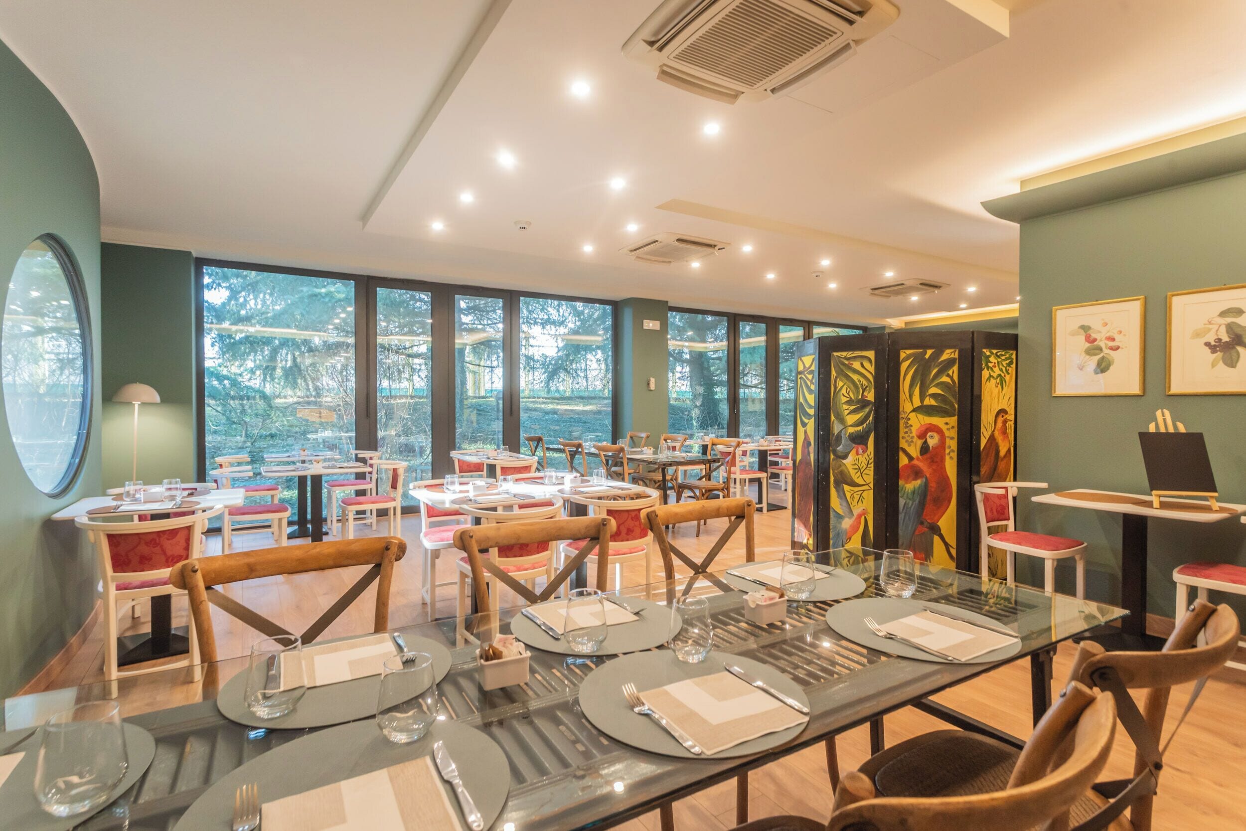 A restaurant with multiple tables set for dining, large windows offering a view of trees, and a ceiling with recessed lighting.