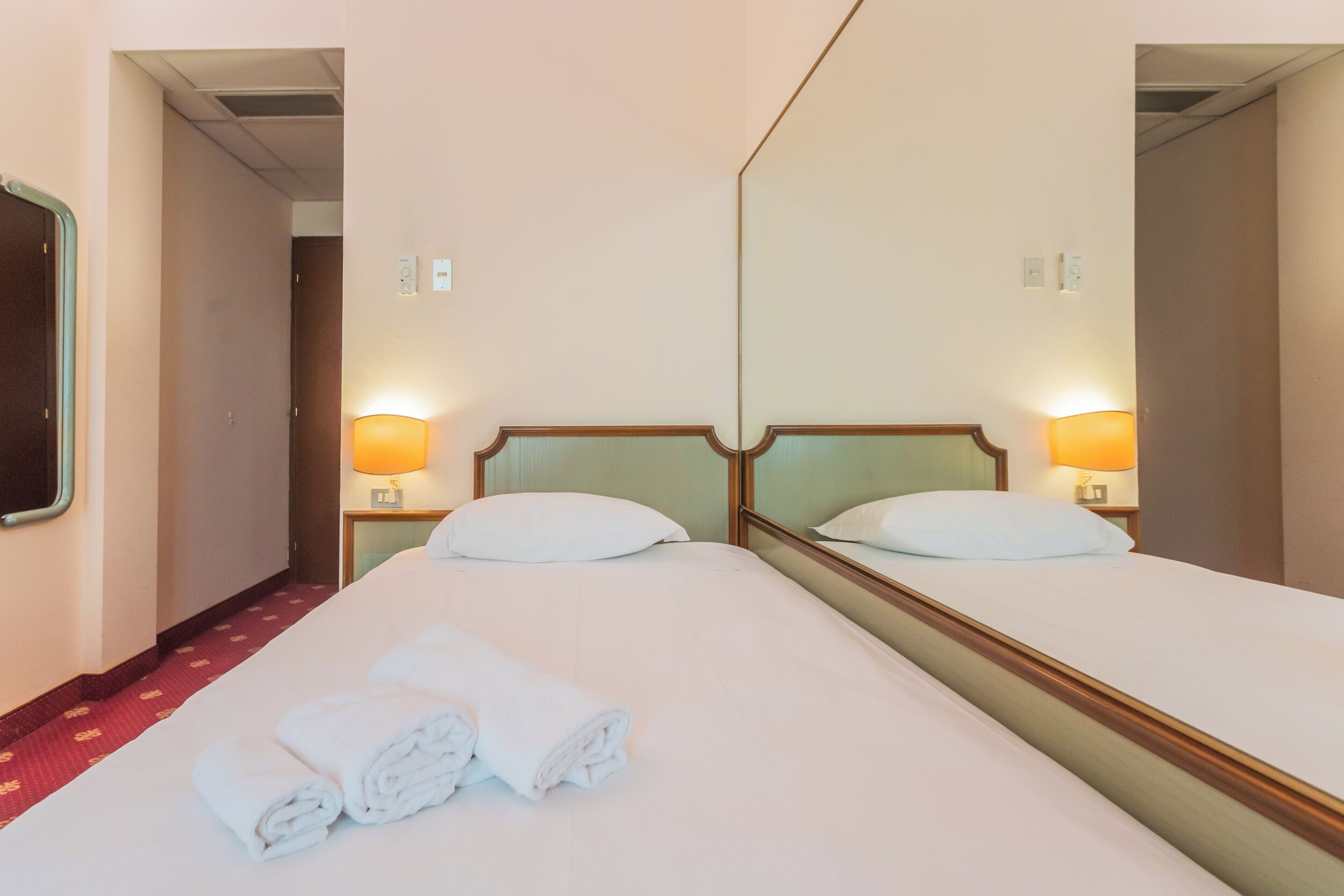 A hotel room with a large bed, two bedside lamps, a mirror, and a neatly folded towel on the bed.