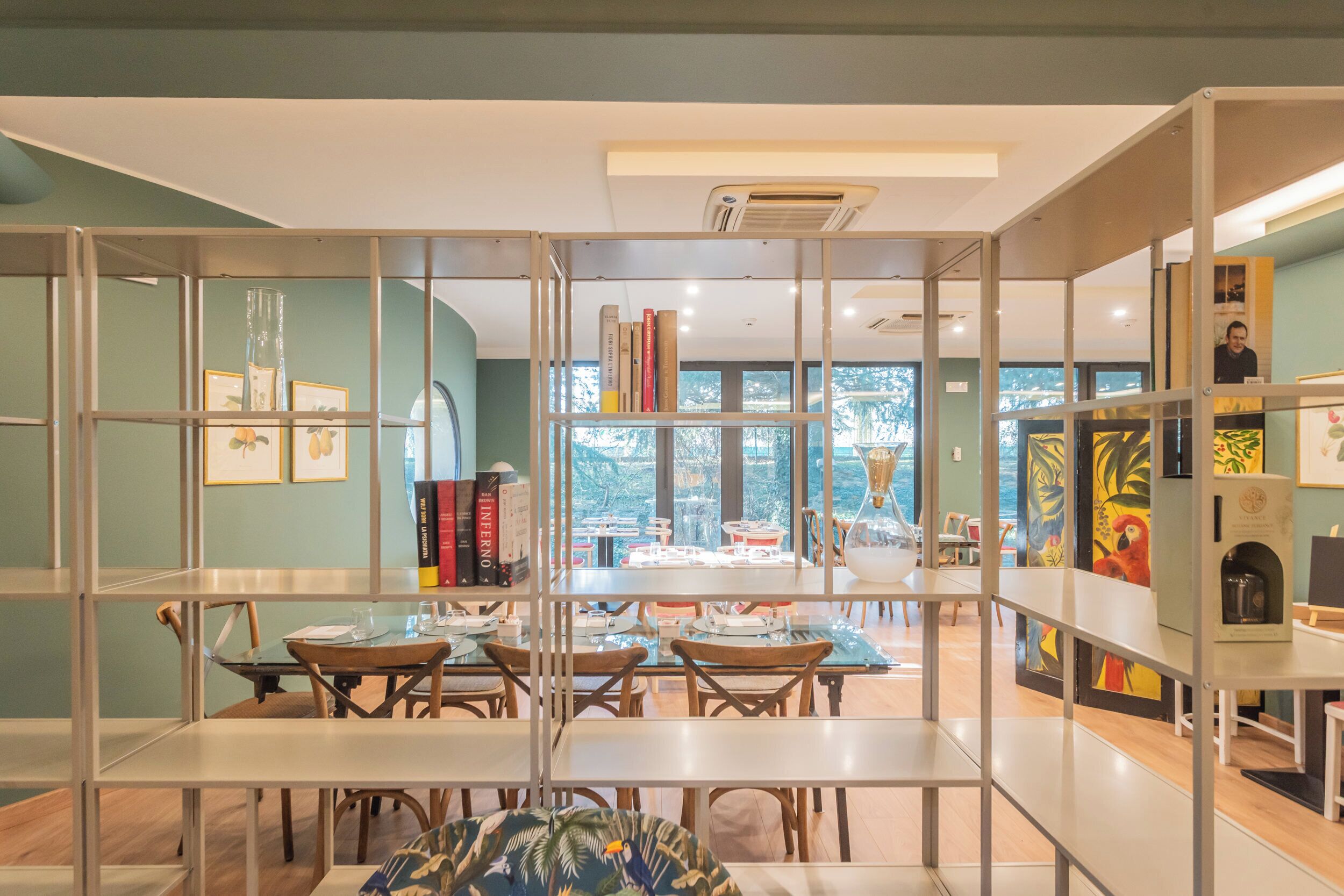 Un comedor moderno con divisiones de vidrio, mesas y sillas de madera. Hay estanterías con libros e imágenes enmarcadas. La habitación tiene ventanas amplias con vistas a árboles y edificios.