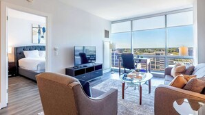 Ein modernes Hotelzimmer mit Bett, Fernseher, Sitzecke und großen Fenstern mit Blick auf die Stadt.