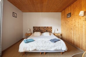 Une chambre avec un plafond en bois, un lit recouvert d’une couverture blanche et moelleuse, deux tables de chevet avec des lampes, et un panneau mural en bois.