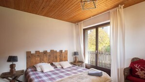 Une chambre avec une tête de lit en bois, une grande fenêtre avec des rideaux, un lit recouvert d’une couverture à carreaux et un fauteuil en osier agrémenté d’un coussin rouge.