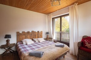 Een slaapkamer met een houten hoofdbord, een groot raam met gordijnen, een bed met een geruit sprei en een rieten stoel met een rode kussens.