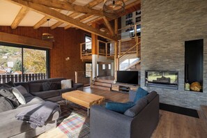 Un vaste espace de vie comprenant un grand canapé, une table basse et un téléviseur. On y trouve également une cheminée et un balcon avec vue sur l’extérieur.
