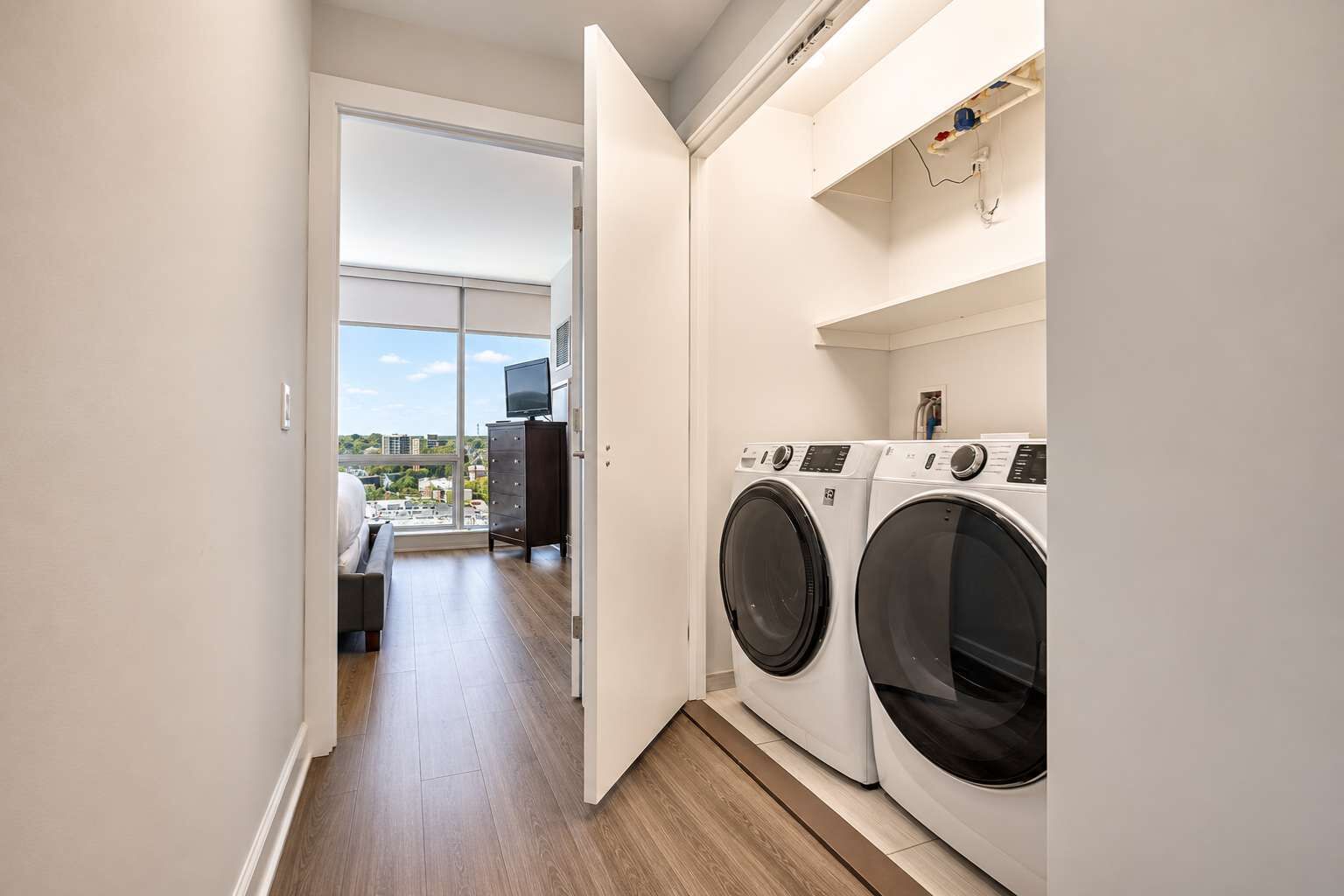 A laundry room with two front-loading washing machines, a television on a dresser, and a view of the city through a large window.