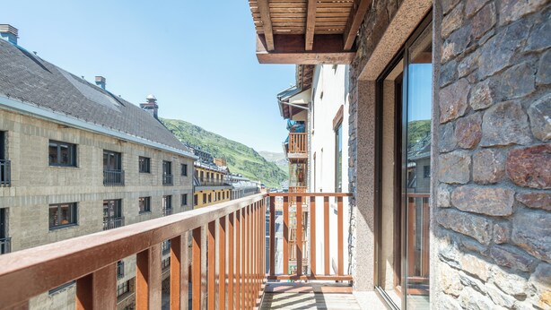 Un balcon doté d’une balustrade en bois, donnant sur une rue bordée d’immeubles et offrant une vue sur un paysage vallonné.