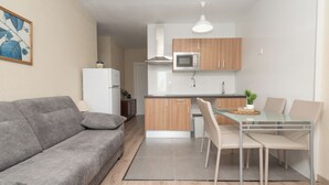 Un espace de vie compact comprenant un coin cuisine, un espace repas avec une table et des chaises en verre, et un canapé gris.
