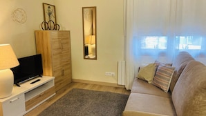 Un salon comprenant un téléviseur, un meuble en bois, un miroir et un canapé avec des coussins.