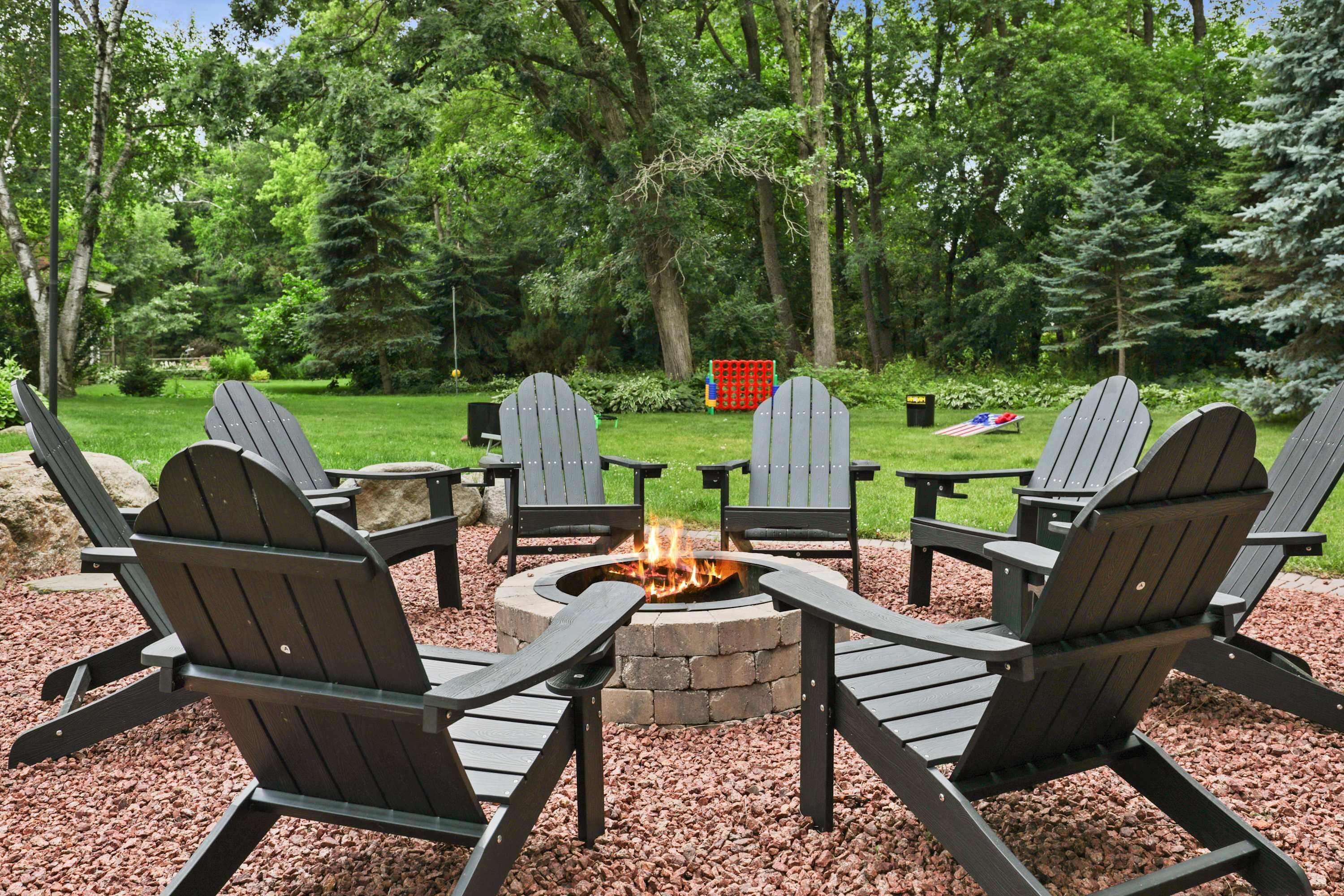 A circular fire pit surrounded by Adirondack chairs in a wooded outdoor area.