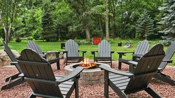 A circular fire pit surrounded by Adirondack chairs in a wooded outdoor area.