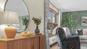Een gezellige woonkamer met een houten kast, een lamp, een vaas met planten, een grijze fauteuil en een groot raam.