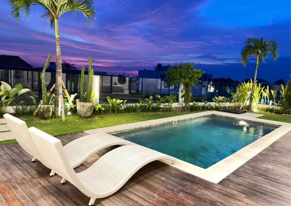 A rectangular pool with lounge chairs on a wooden deck, surrounded by tropical plants and palm trees, with a view of buildings in the background. - The R Villa Ranakah (Mengwi)