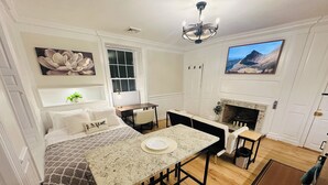 A compact, well-lit room with a bed, a dining table, a fireplace, and a large window.