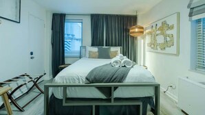 Una camera da letto con un letto, una ventola a soffitto, una finestra con tende, un comodino e un'opera d'arte appesa al muro.