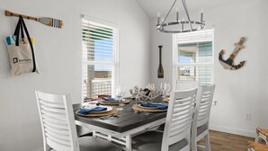 A dining area with a dark wooden table set for four, white chairs, and a chandelier. - Green Casita, Heated Pool, Fire Pit & Outdoor Bar (Port Aransas)