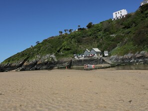A beach with a white building on a hill, a sandy shore, and a small dock with boats. - Chynoweth (Newquay)