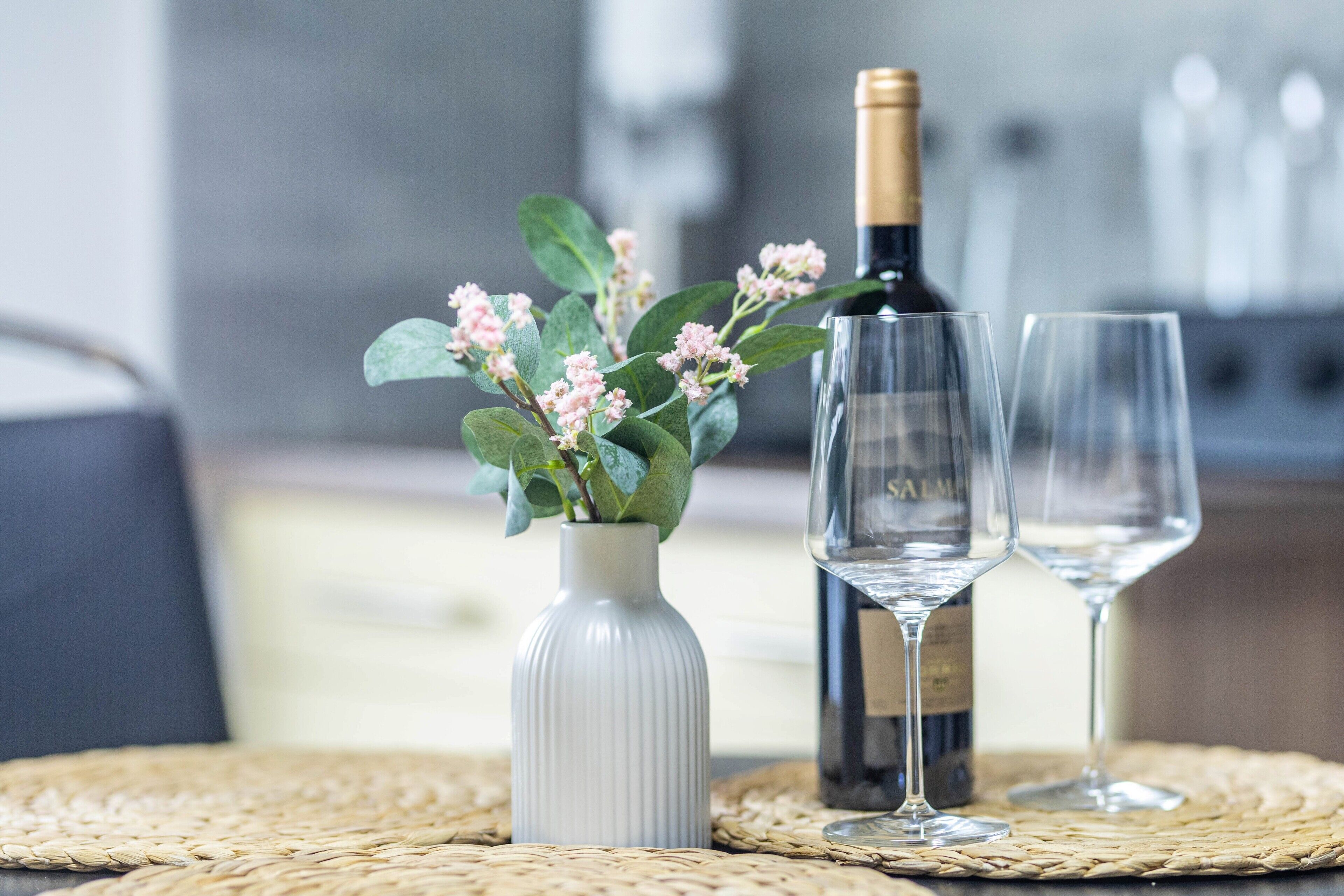 Una botella de vino, dos copas y un jarrón con flores sobre una base tejida.